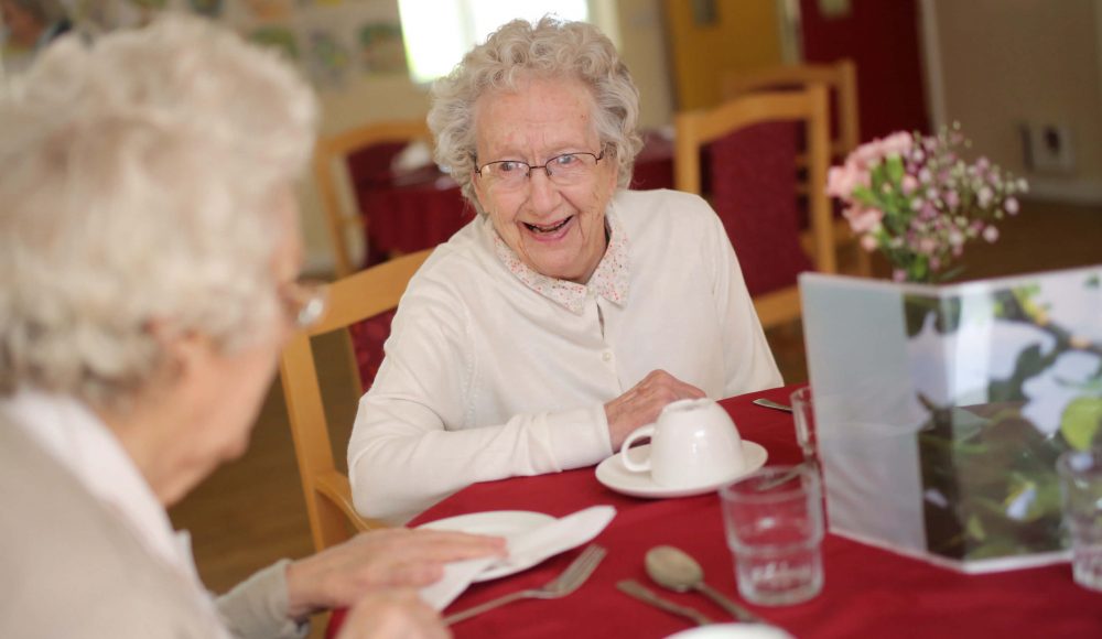 Two residents talk over a cup of tea at the dining table in a care homes dining room